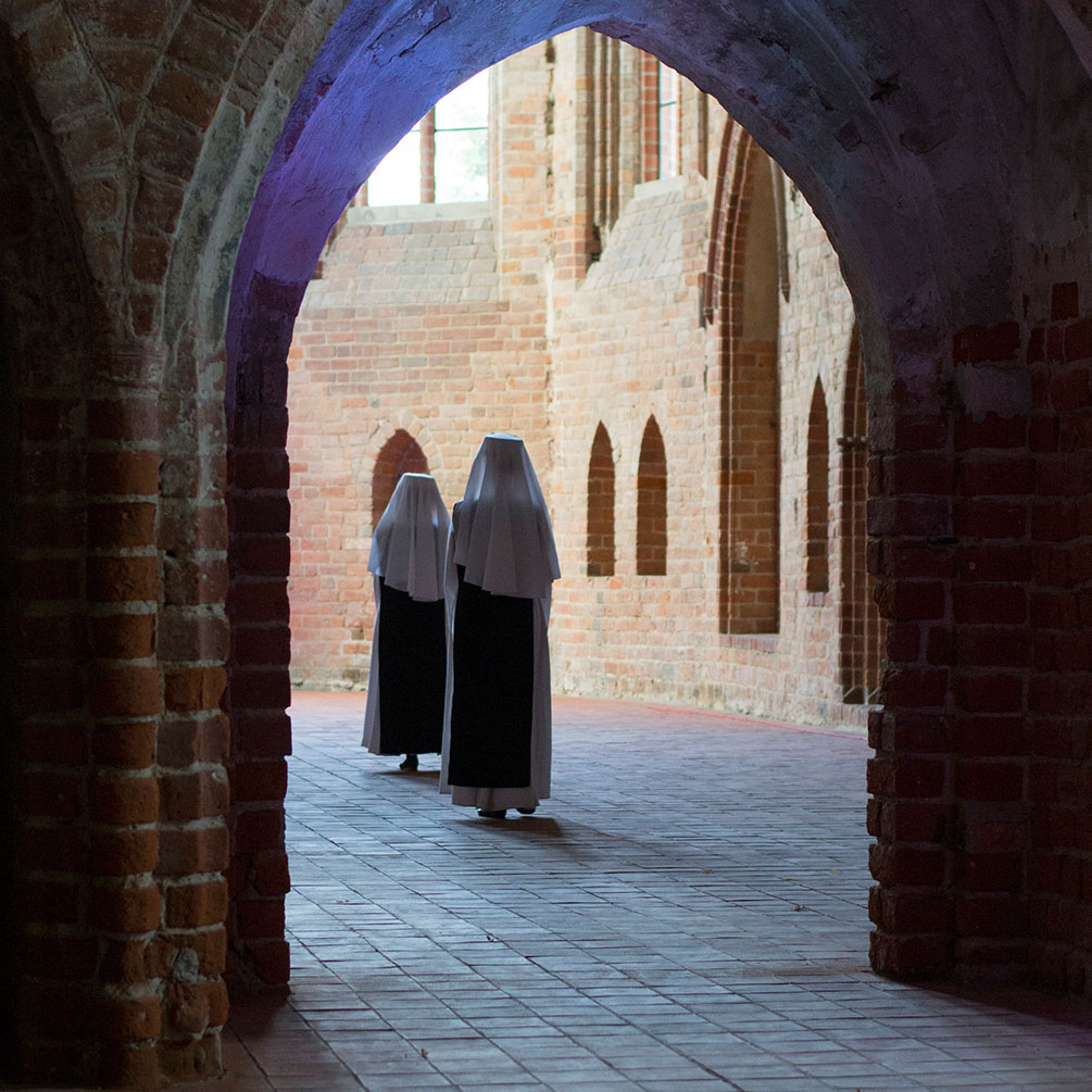 Zwei Nonnen stehen im Chorraum. Sich sind von hinten zu sehen. Im Vordergrund der Torbogen der den Chorraum vom Hauptraum der Kirche trennt.